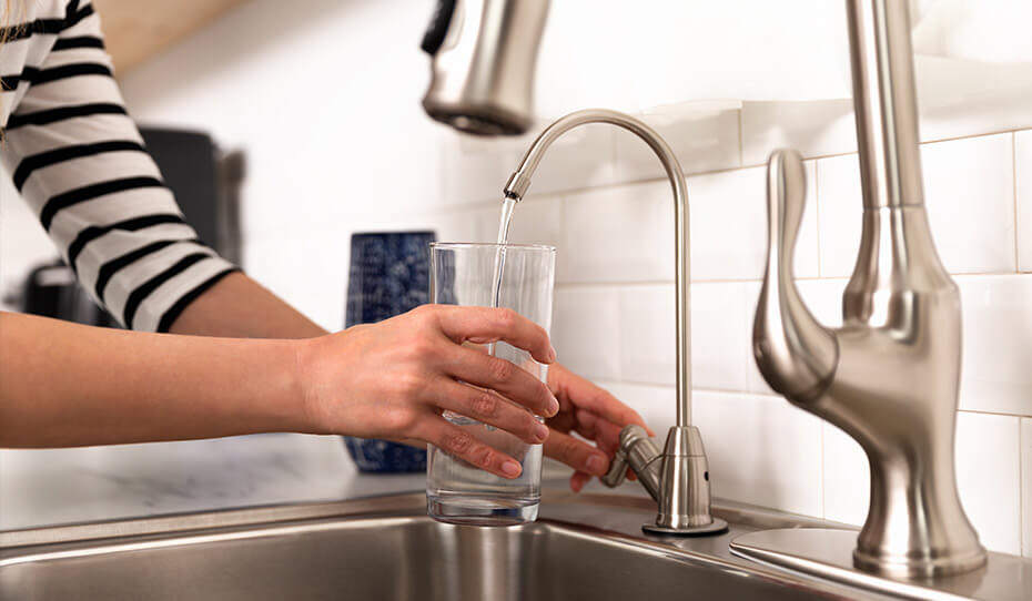 A woman is filling a glass of water from a kitchen sink with a water purifier.
