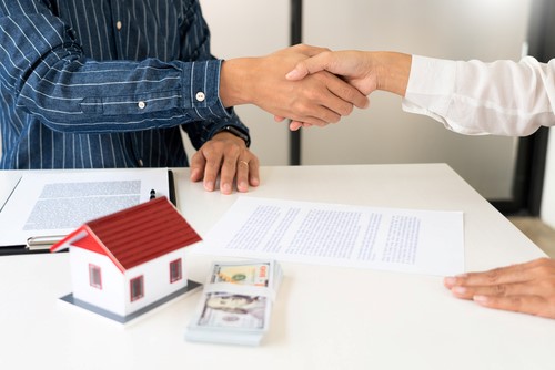 Two people shaking hands in front of a house model, symbolizing property to buy.