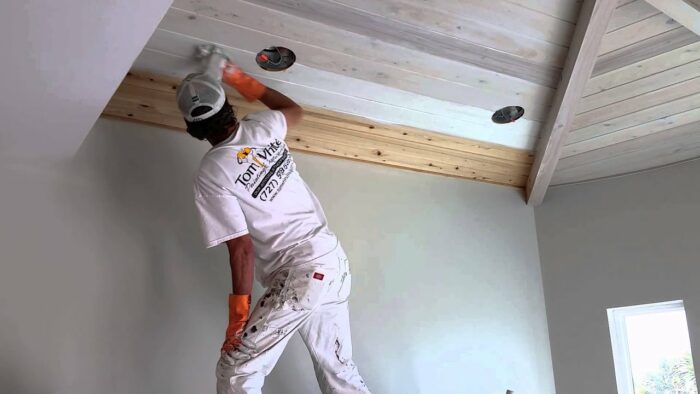 A man is painting a tongue and groove ceiling in a room.