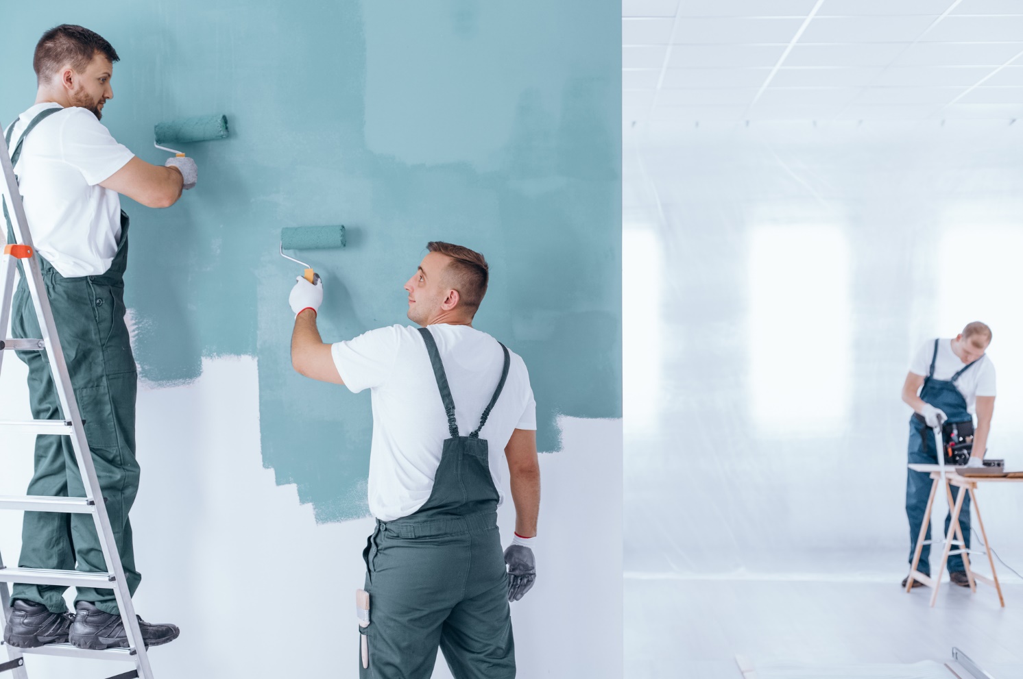 Three men working for a painting company paint a wall in a room.