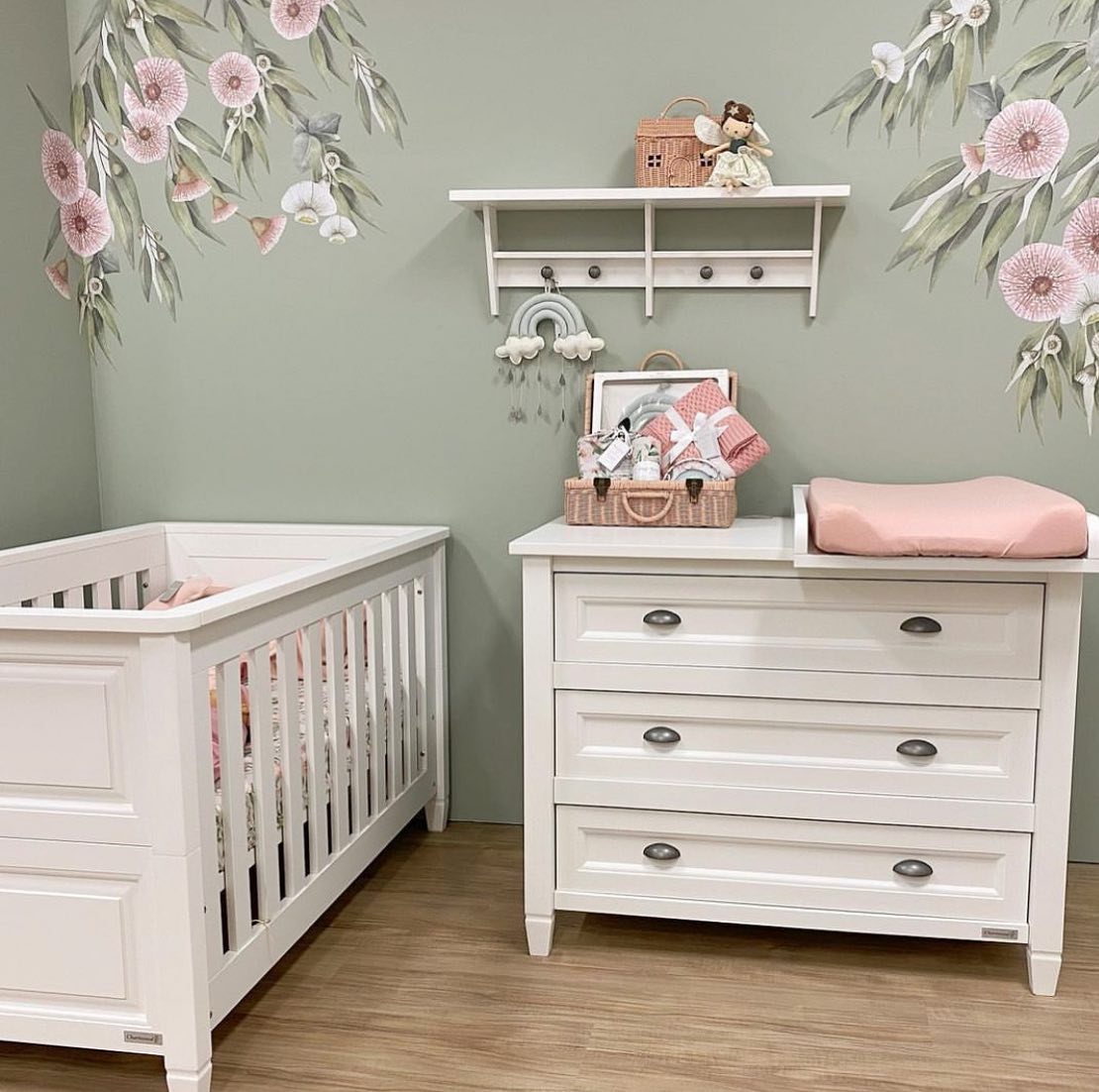 A sage green nursery with a crib, dresser, and a wall mural.