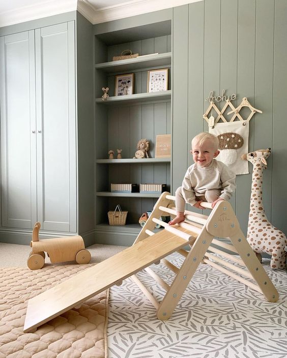 A baby is playing on a wooden slide in a sage green nursery.