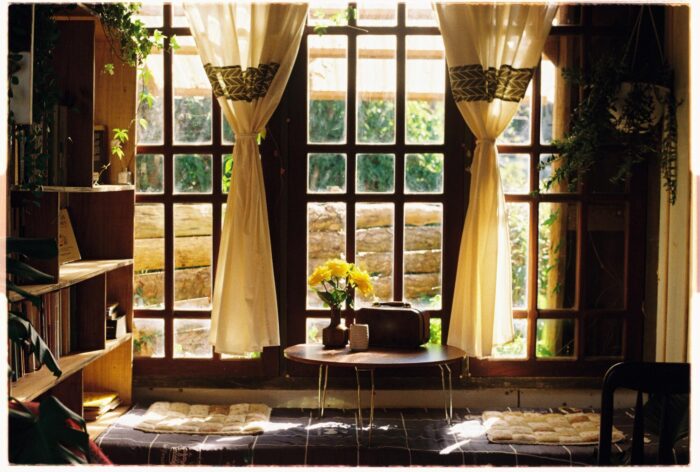 Large grid-style windows with cream curtains, indoor plants, and warm natural light in a cozy living space