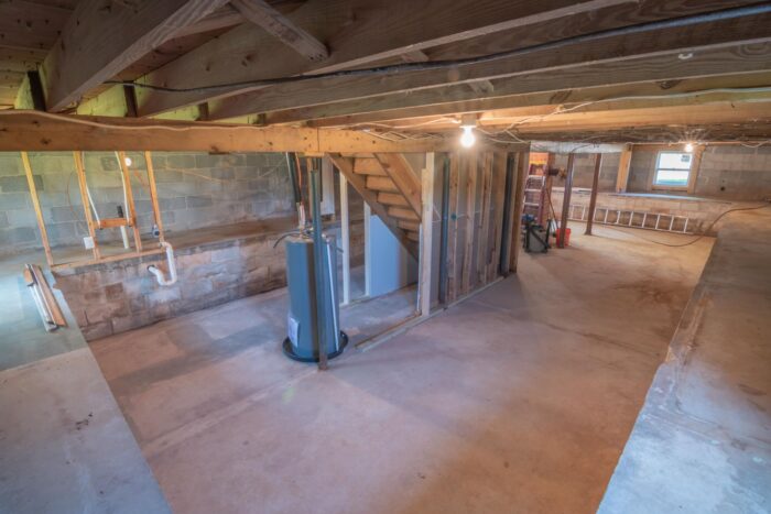 Unfinished basement with exposed wooden ceiling beams, concrete block walls, a water heater, plumbing, and a staircase before renovation.