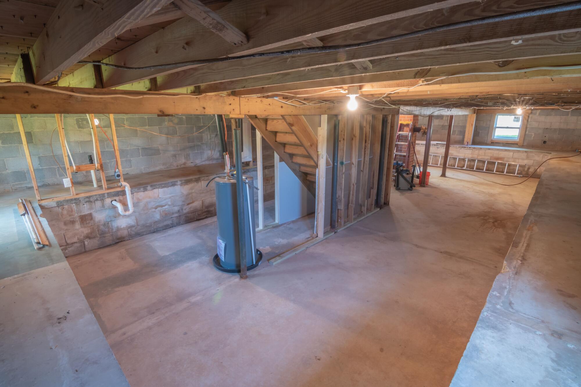 Unfinished basement with exposed wooden ceiling beams, concrete block walls, a water heater, plumbing, and a staircase before renovation.