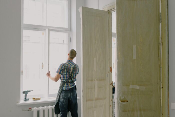 Home renovation in progress with a worker inspecting a large window, protected interior doors, and tools on a windowsill.
