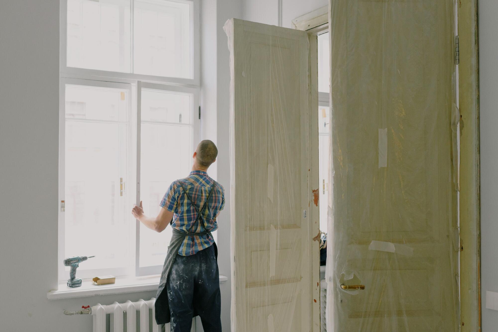Home renovation in progress with a worker inspecting a large window, protected interior doors, and tools on a windowsill.