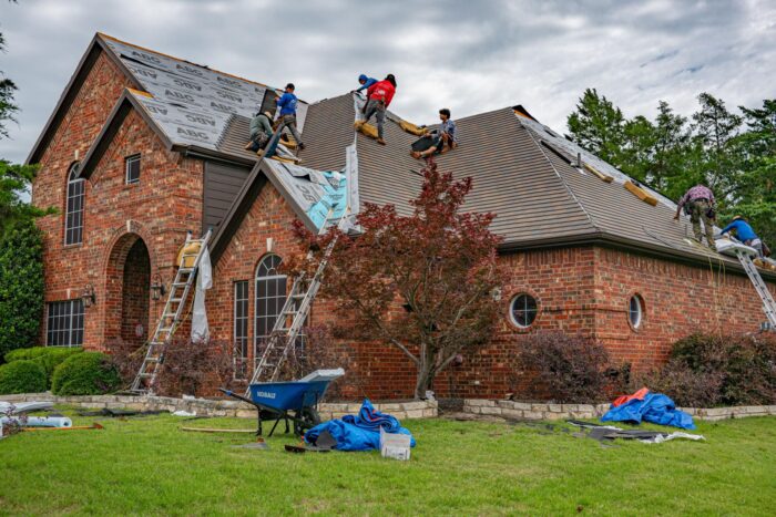 Roofers repairing and replacing sections of a brown shingle roof on a two-story red-brick house.